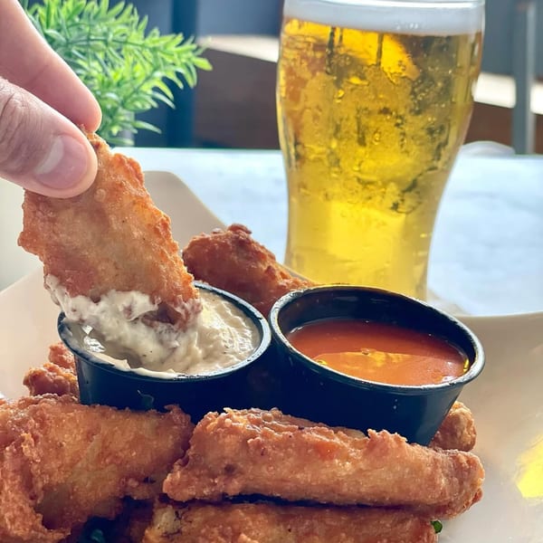 A hand dips a crispy fried chicken strip into a white sauce, with a beer in the background.