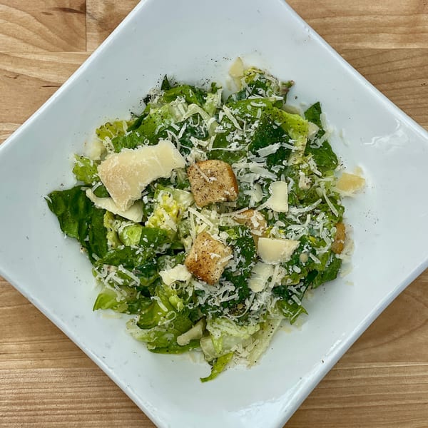 A Caesar salad with romaine lettuce, croutons, and parmesan cheese in a white square bowl.